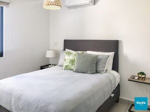 A neatly arranged bedroom with a bed, a bedside table with a lamp, a small shelf with a plant, and an air conditioning unit on the wall.