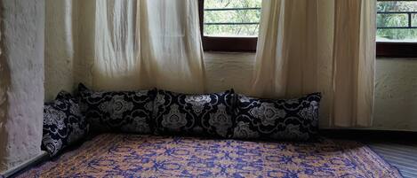 A room with a patterned bedspread, a window with curtains, and a view of greenery outside.