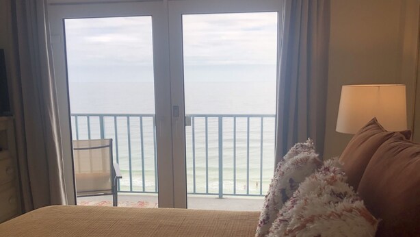 Una camera d'albergo con un letto grande, vista sul mare attraverso una porta scorrevole, una sedia vicino alla porta e una lampada sul comodino.