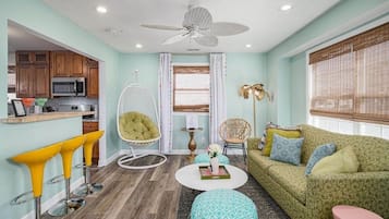A living room with a green sofa, yellow bar stools, a white ceiling fan, and a kitchen area with wooden cabinets and a microwave.