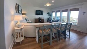 A modern living room with a sofa, a small side table, a ceiling fan, and a dining area with chairs. - Thibodeaux's Beach House (Grand Isle)
