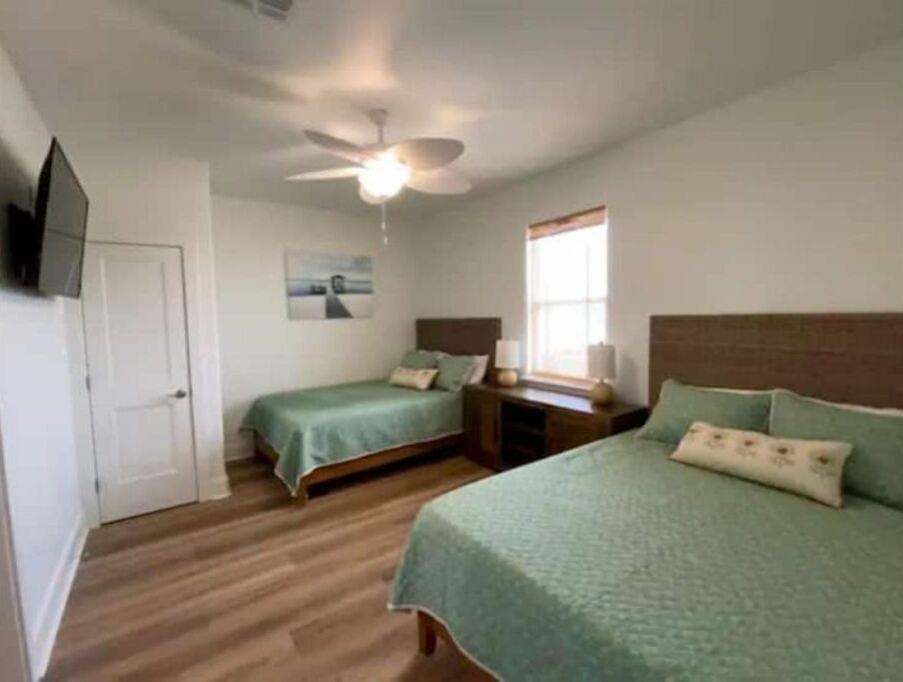 A bedroom with two single beds, a ceiling fan, a window with curtains, and a flat-screen TV mounted on the wall.