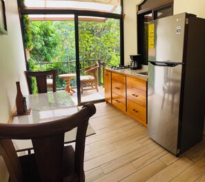 A kitchen with a refrigerator, wooden cabinets, a table with chairs, and a view of greenery outside. - Enchanted Sloth (Manuel Antonio)