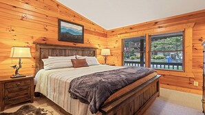 A wooden-paneled bedroom with a bed, two bedside tables, lamps, a painting, and a window with blinds.