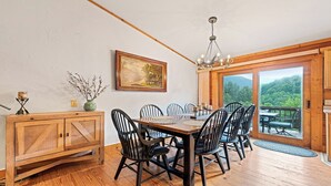 A dining area with a wooden table and chairs, a chandelier, a painting on the wall, and a view of a forest through a glass door.