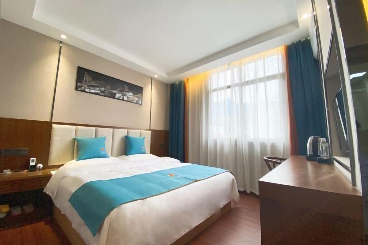 A hotel room with a bed, a desk, a chair, a painting on the wall, and a window with curtains.