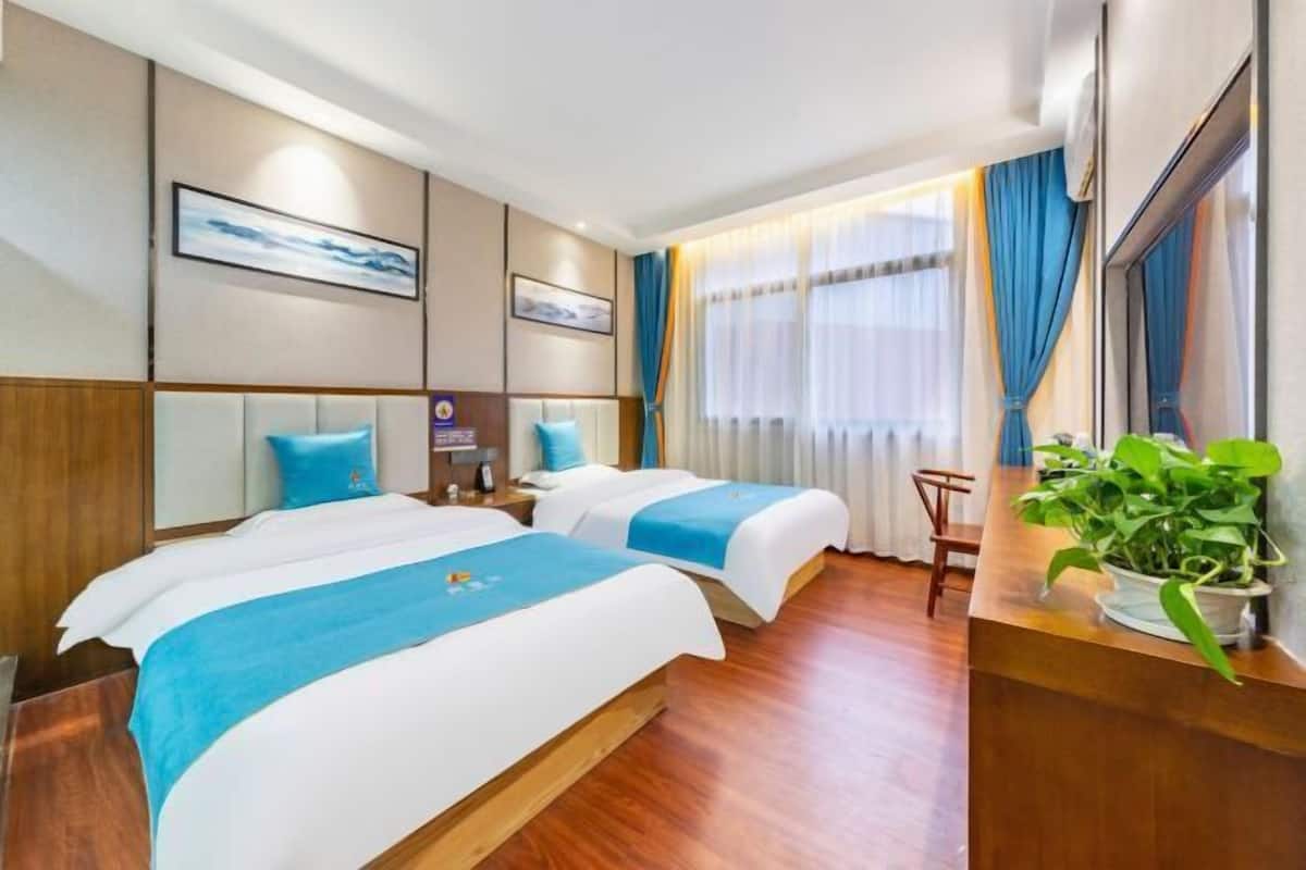 Ein Hotelzimmer mit zwei Betten, einem Holzkopfstück, einem Nachttisch, einer Pflanze und einem Fenster mit Vorhängen.