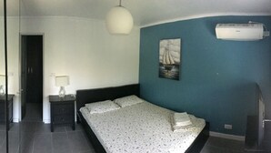 Habitación de hotel con una cama, una mesita de noche con lámpara, aire acondicionado instalado en la pared y un cuadro con barcos de vela.