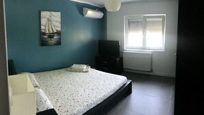 Un dormitorio con cama, televisor, ventana con persianas y un cuadro en la pared.