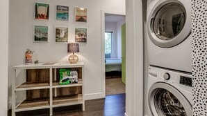 A laundry area with a washing machine, a shelf with travel-themed decor, and a hallway leading to a bedroom. - Jungle House: 2 Bed Condo w/parking & Patio (Denver)