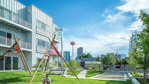 A modern building with a grassy courtyard, swing sets, and a city skyline in the background. - Jungle House: 2 Bed Condo w/parking & Patio (Denver)