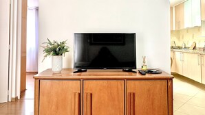 A modern living room with a flat-screen TV on a wooden cabinet, a potted plant, and a kitchen visible in the background. - Apartamento Próx Tiete Plaza Shopping (São Paulo)