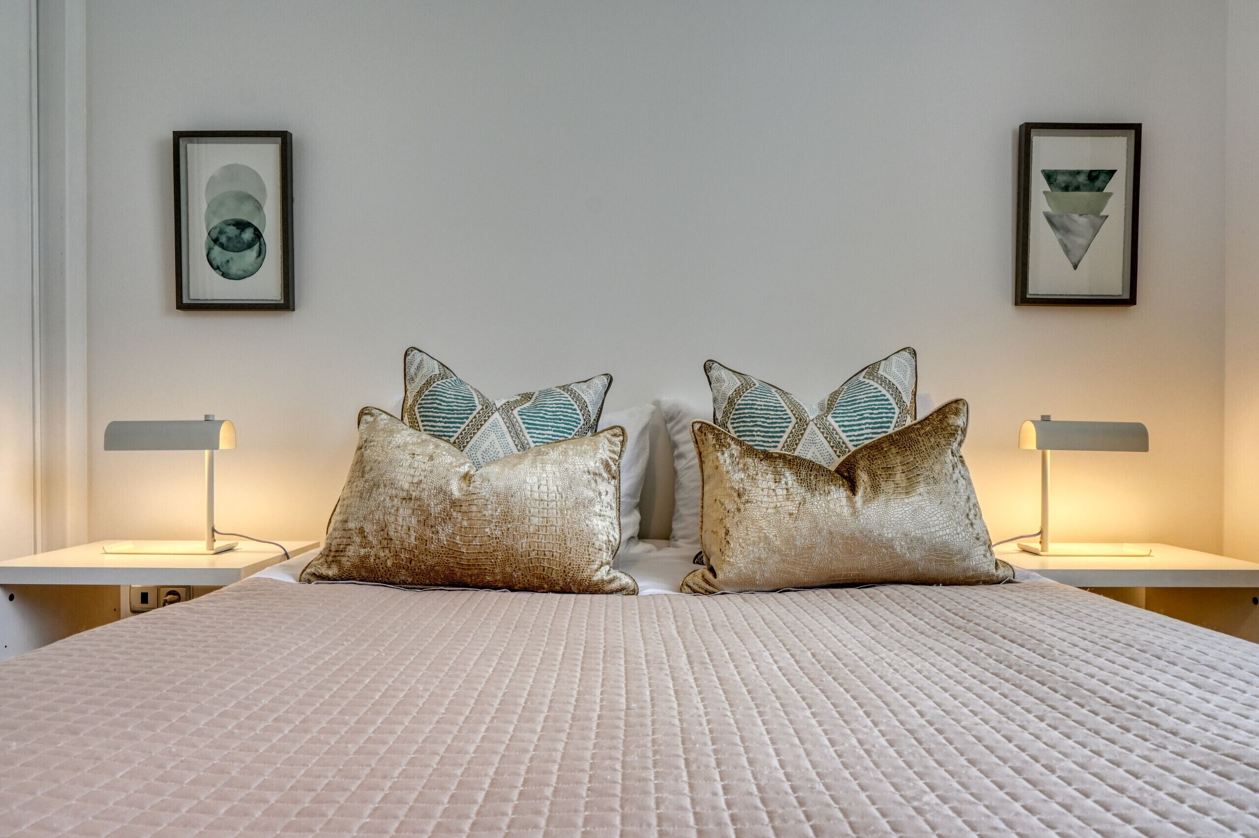 Una cama bien tendida con una colcha blanca texturizada, flanqueada por dos mesitas de noche con lámparas, cada una con un cuadro enmarcado.