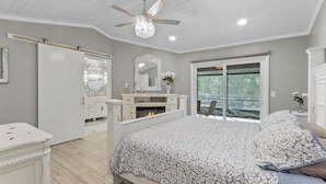 A bedroom with a large bed, a ceiling fan, a fireplace, and a window with a view.