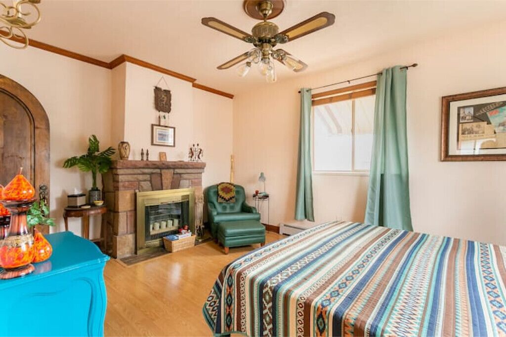 A room with a fireplace, a bed with a patterned bedspread, a ceiling fan, and a window with curtains.