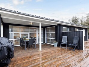 Um alpendre coberto com um deck de madeira, uma área de refeições com cadeiras e uma mesa, e uma parede externa preta com janelas.