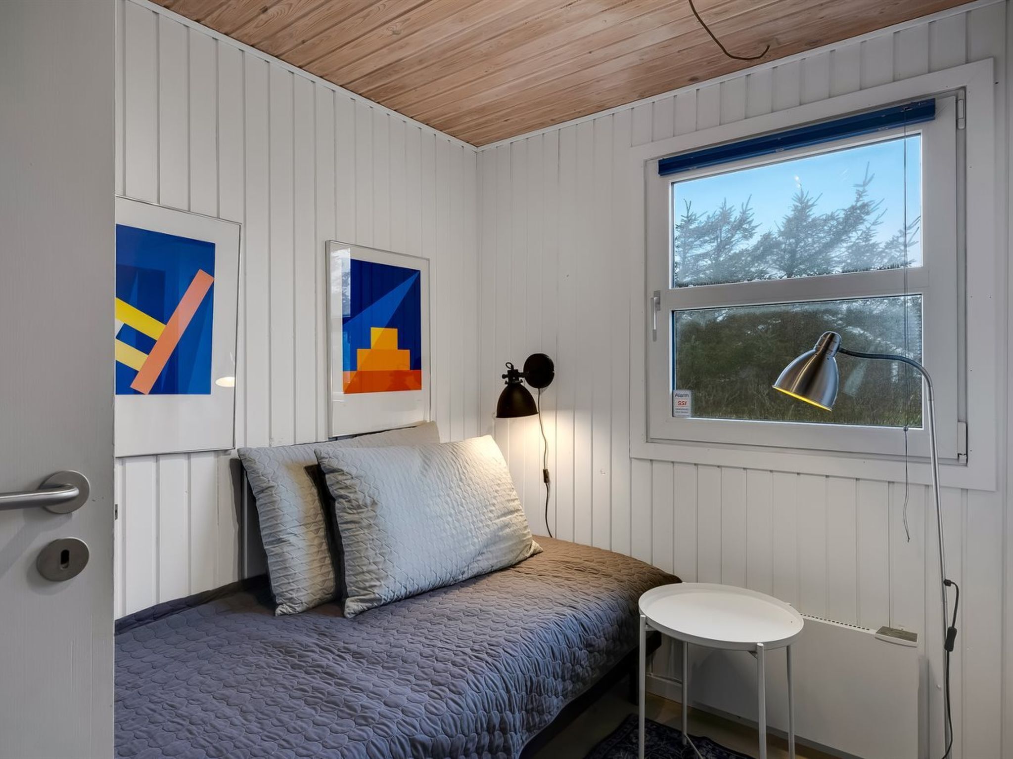 Une chambre à coucher avec un lit, une petite table, une lampe et deux tableaux abstraits accrochés au mur.