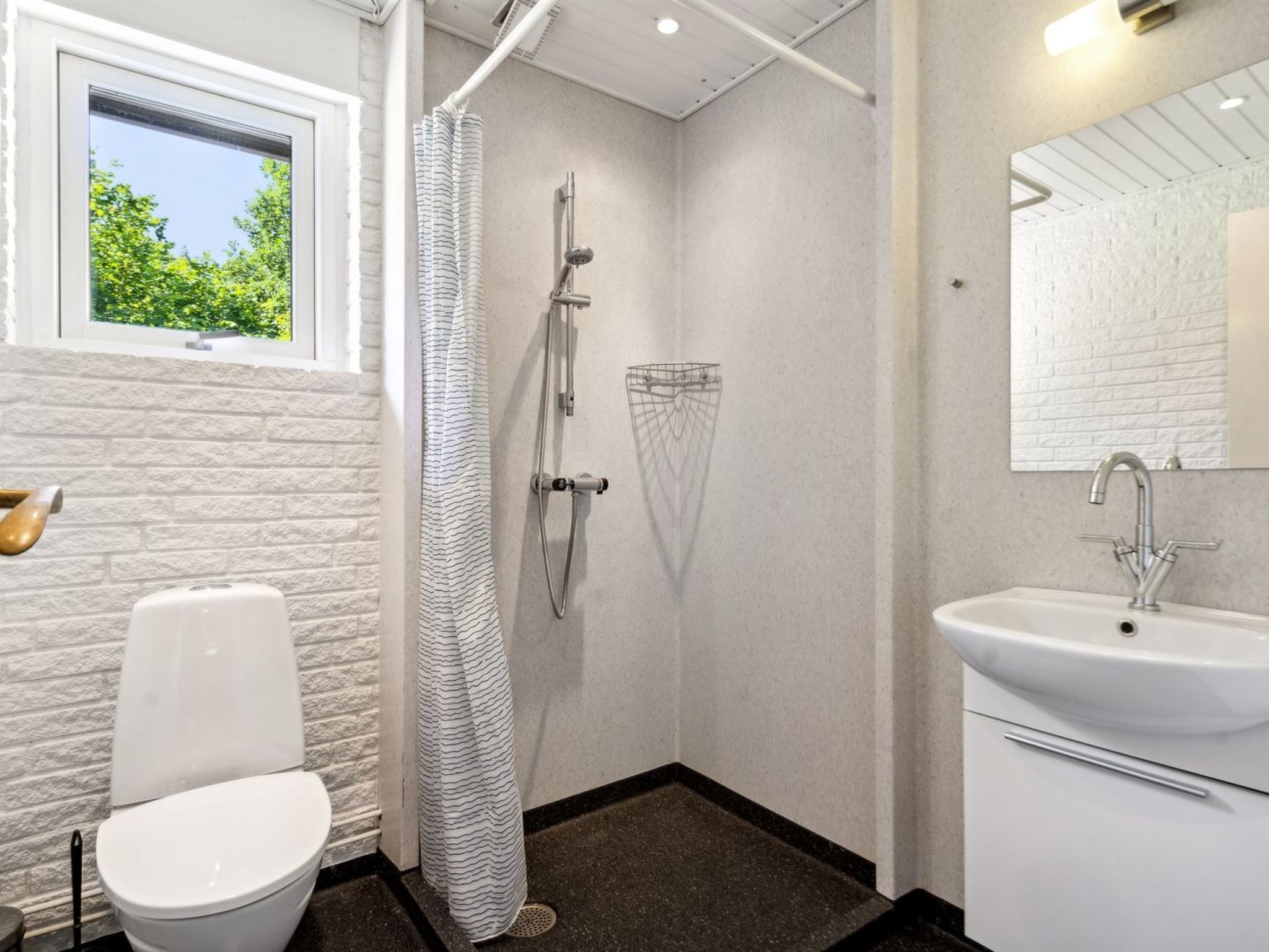 Et badeværelse med toilet, brusebad med badeforhæng, håndvask med spejl og et vindue med udsigt over grønne omgivelser.