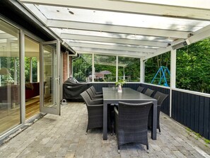 Une terrasse couverte avec une table et des chaises, un barbecue et, au fond, un ensemble de jeux pour enfants avec une balançoire.