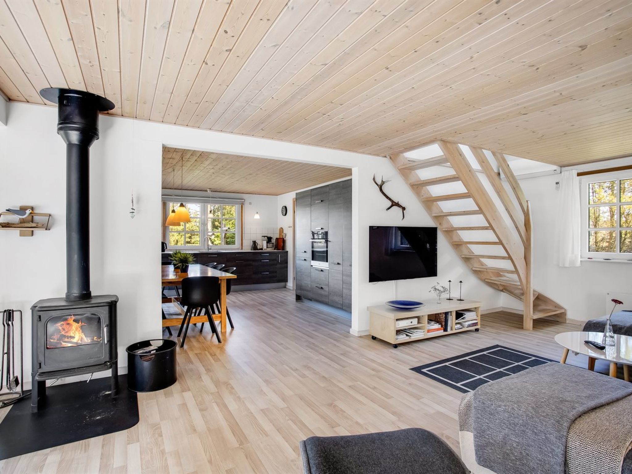 A modern living room with a wood-burning stove, a dining area, a kitchen, and a staircase.