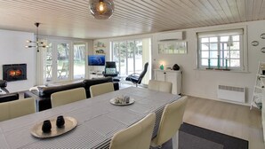 Un salon moderne avec une cheminée, un coin repas avec une nappe à rayures et de grandes fenêtres donnant sur l’extérieur.