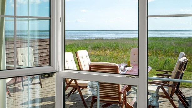 Et værelse med store vinduer med udsigt over en strand. Der er flere stole og et bord med et lys og et glas.