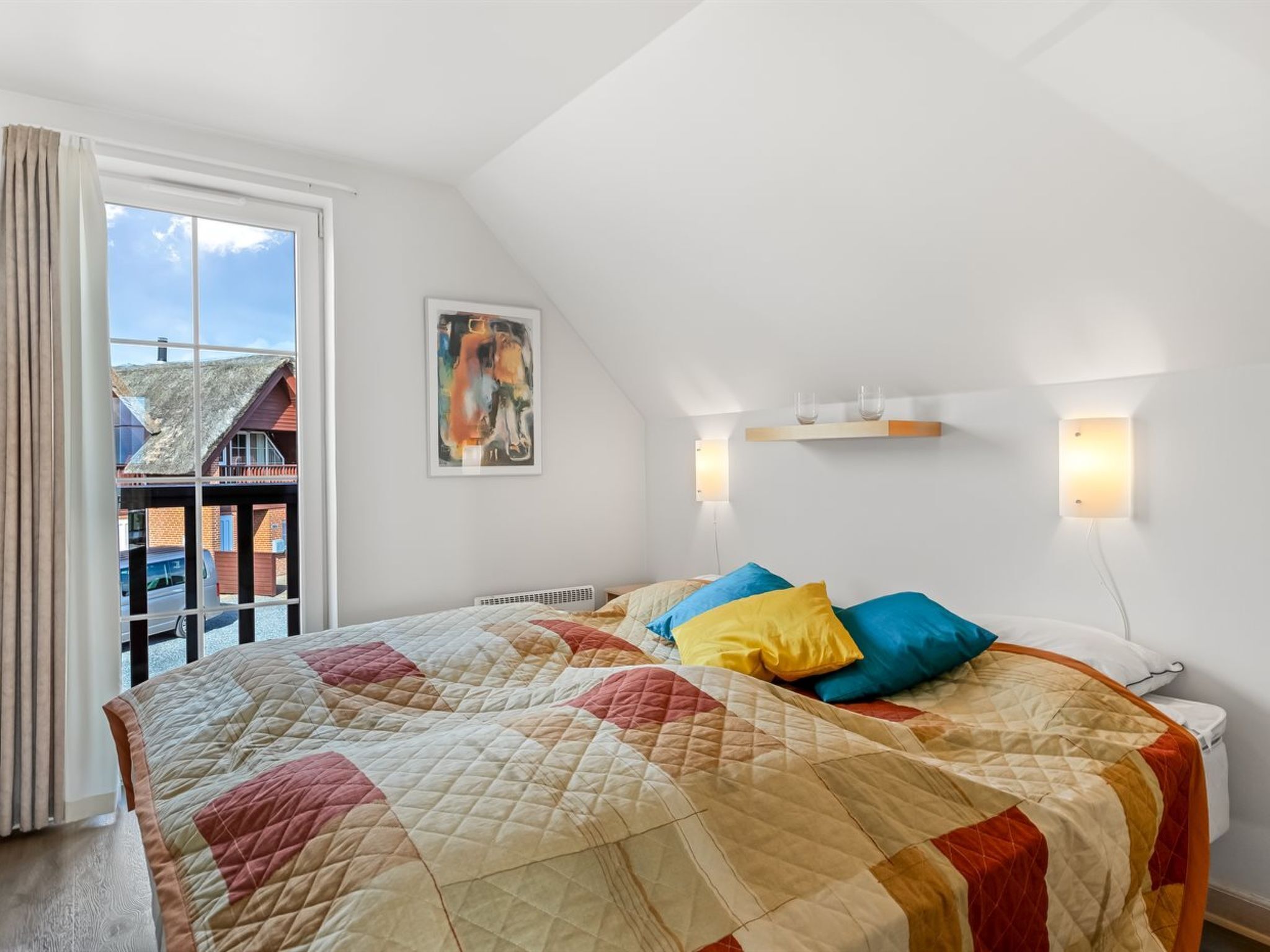 A bedroom with a large bed, a window with curtains, a painting on the wall, and a small shelf with glasses.