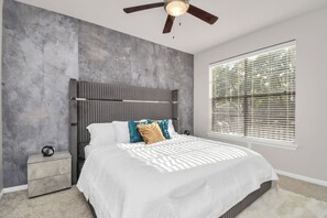 Ein Schlafzimmer mit Bett, Nachttisch, Deckenventilator und Fenster mit Jalousien.