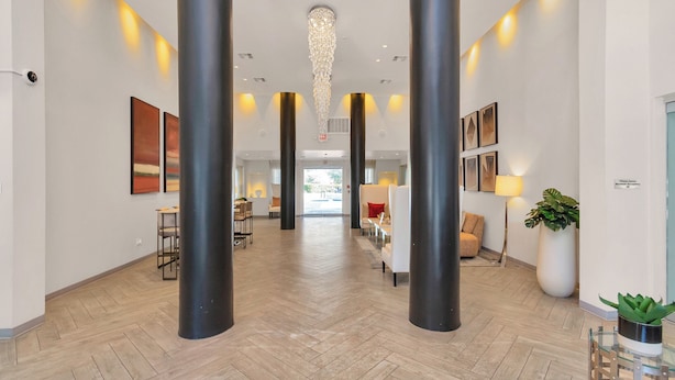 Um hall amplo com padrão de piso em herringbone, colunas pretas e um lustre central.