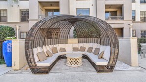 Uma área de estar externa com design curvo, incluindo uma mesa redonda, cercada por uma estrutura de vime, em frente a um edifício com vários andares.