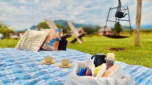 Un'area picnic su una coperta a righe blu e bianche, con un sacco di cibo, due tazze e un bollitore, in un prato con altalene sullo sfondo.
