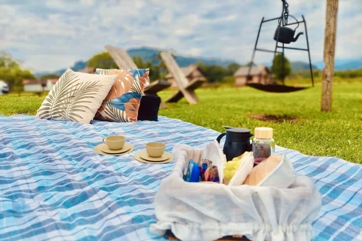 A picnic setup on a blue and white striped blanket with a bag of food, two cups, and a kettle on a grassy field with a swing set in the background.