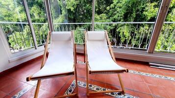 Two wooden lounge chairs with white cushions on a tiled floor, positioned in front of large windows with a view of green trees.