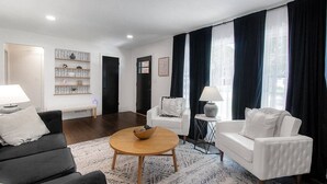 Uma sala de estar moderna com um sofá, duas poltronas, uma mesa de centro e uma mesinha de apoio com uma luminária. Há uma estante com prateleiras estampadas e um quadro na parede.