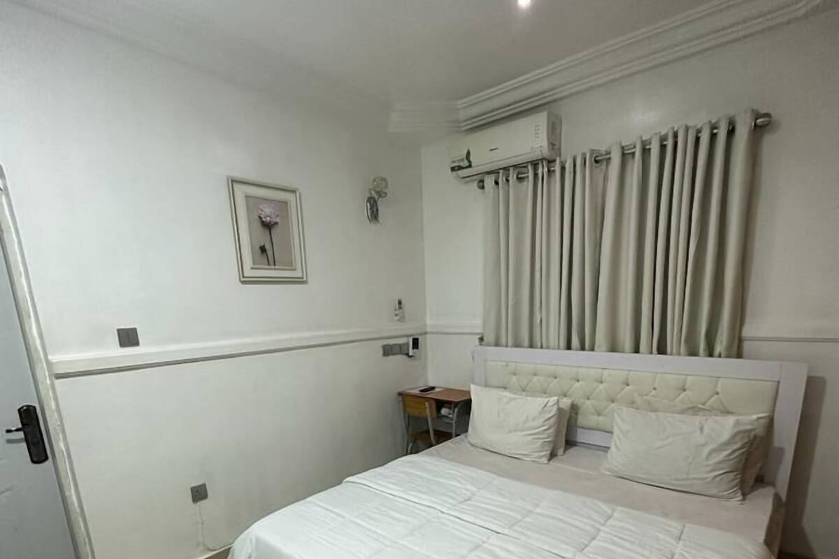 A bedroom with a large bed, a small bedside table, a wall-mounted air conditioner, and a door.