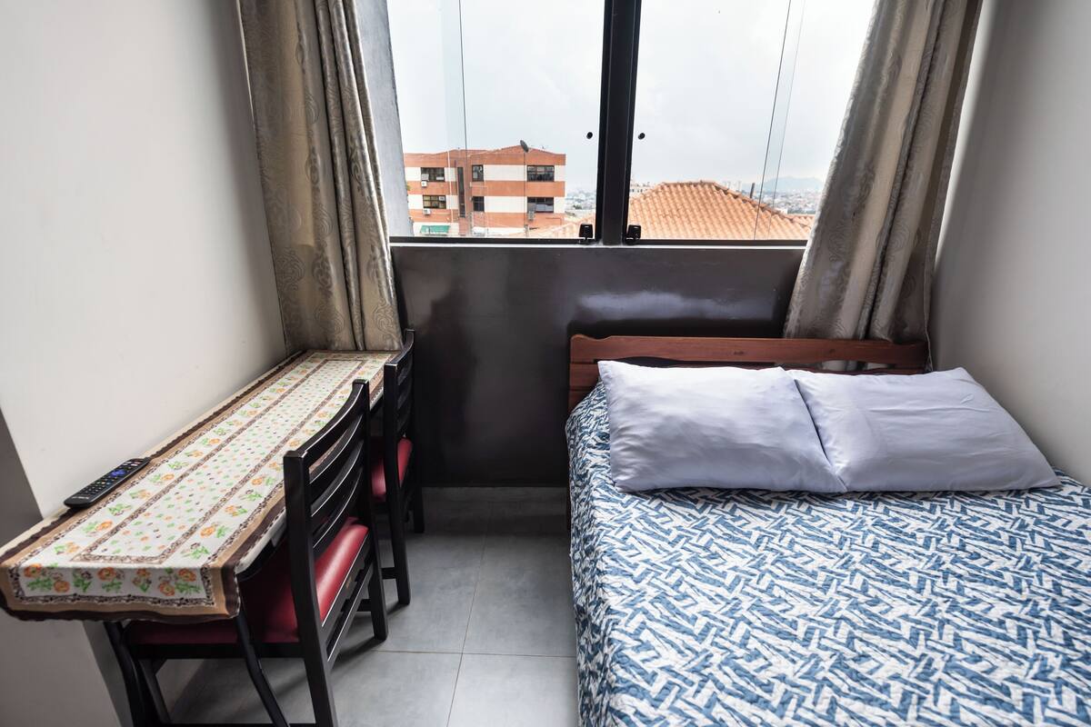 Piccola camera funzionale con un letto, una scrivania e una sedia. Una finestra offre vista sugli edifici esterni.