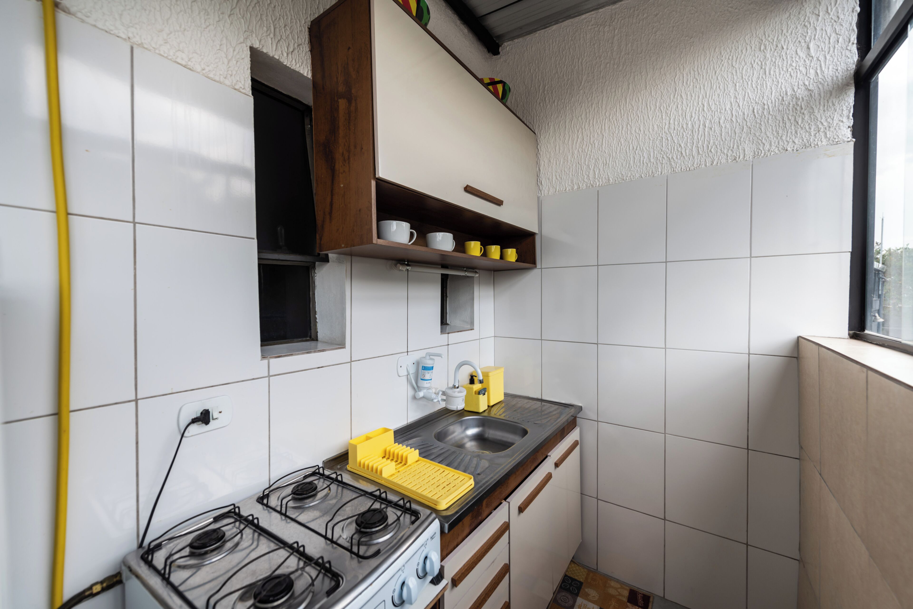 A compact kitchen with white tiles, a gas stove, a sink, and a wooden cabinet above the counter.