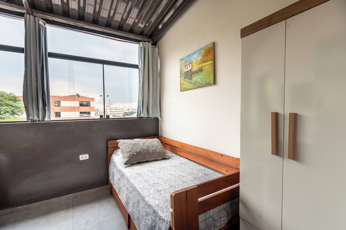 Una camera piccola con un letto singolo, un armadio, una finestra con tende e un quadro al muro.