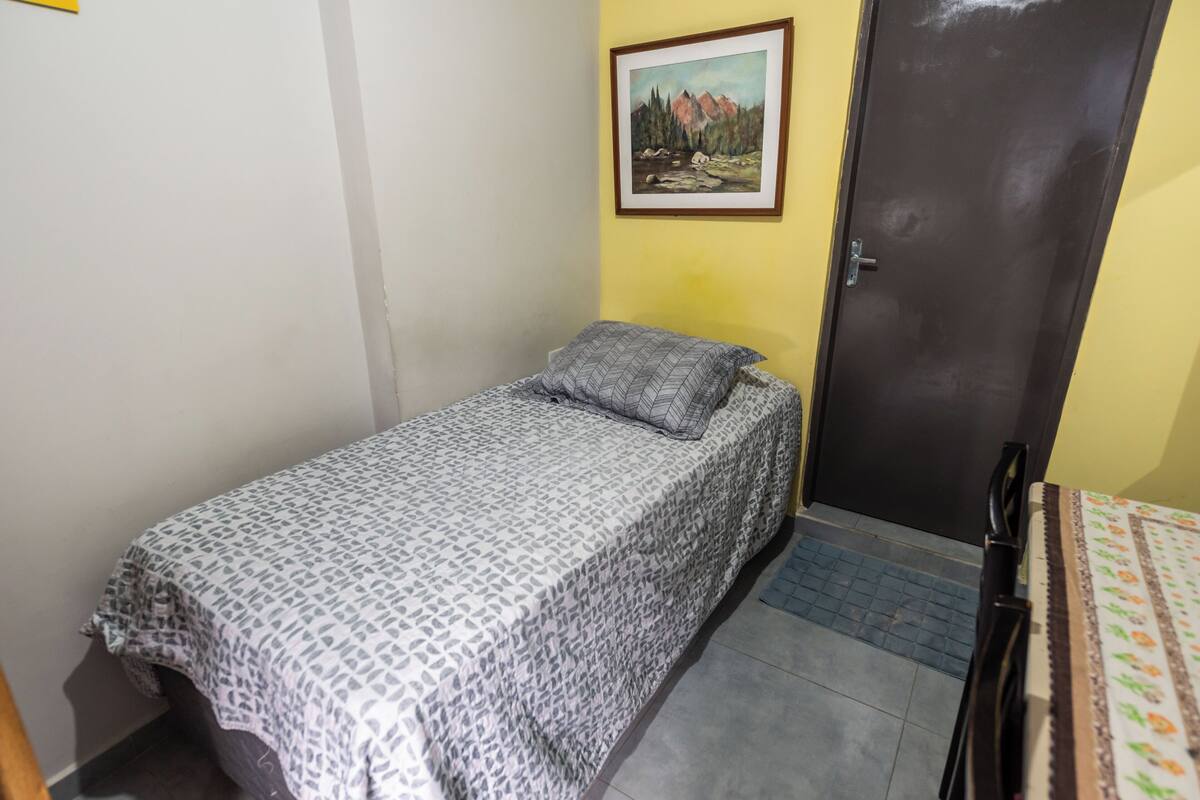 Una camera piccola con un letto singolo, una coperta a motivi, un quadro di un paesaggio appeso alla parete e una porta chiusa.