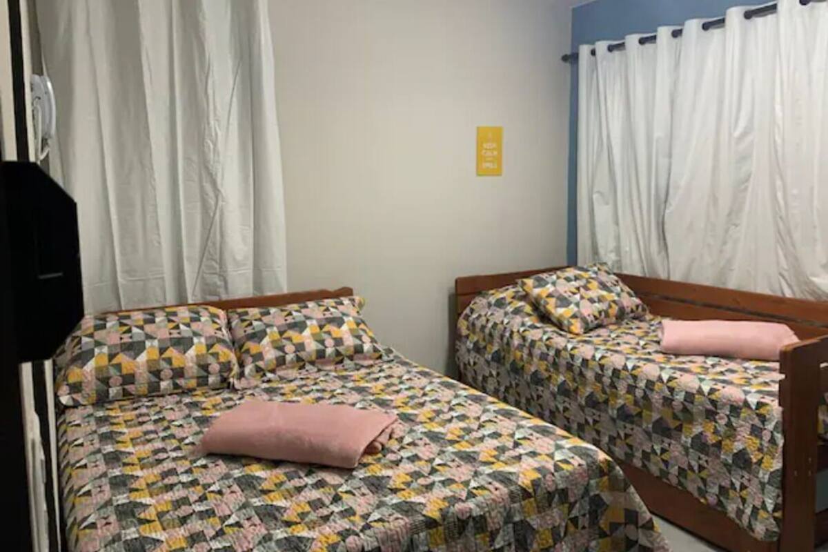 Una stanza con due letti, ognuno con biancheria a motivi decorativi e un cuscino rosa. C'è un cartello giallo sulla parete e tende bianche alle finestre.