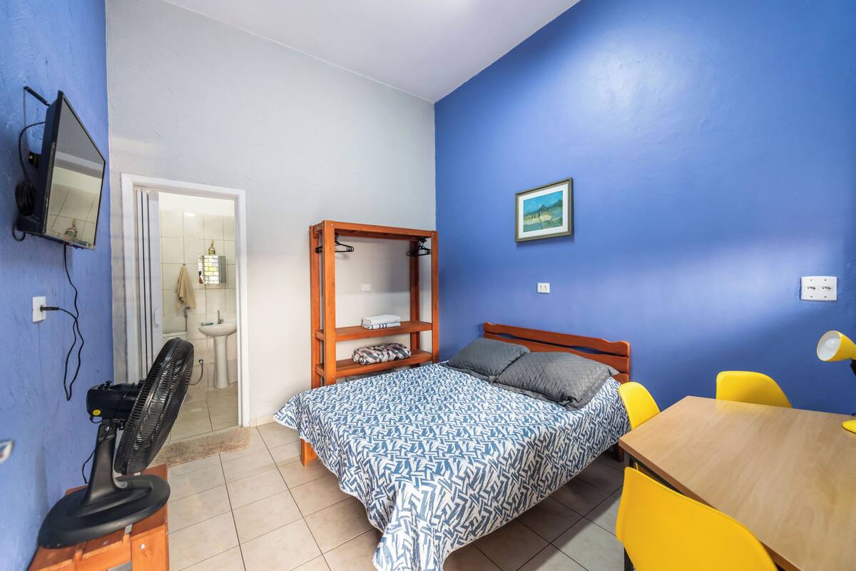 Una camera da letto con un letto, un'armadio in legno, una scrivania con sedia, una ventola e una TV a parete.