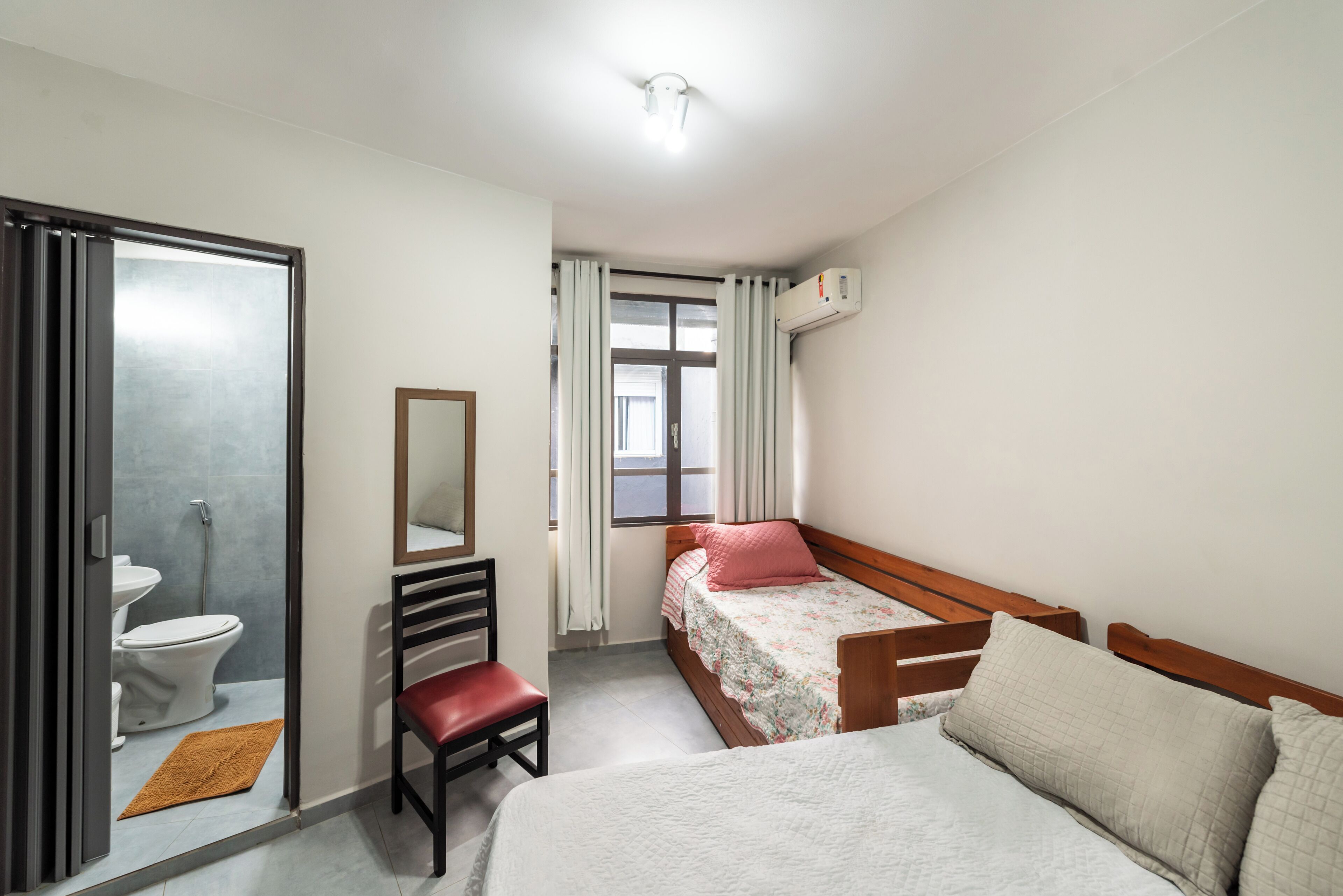 Quarto com cama, cadeira de madeira, mesa de cabeceira, espelho, janela com cortinas e ar condicionado.
