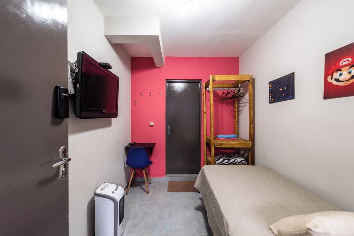 Una stanza piccola e luminosa, con un letto, una scrivania, una sedia, una televisione e una porta.