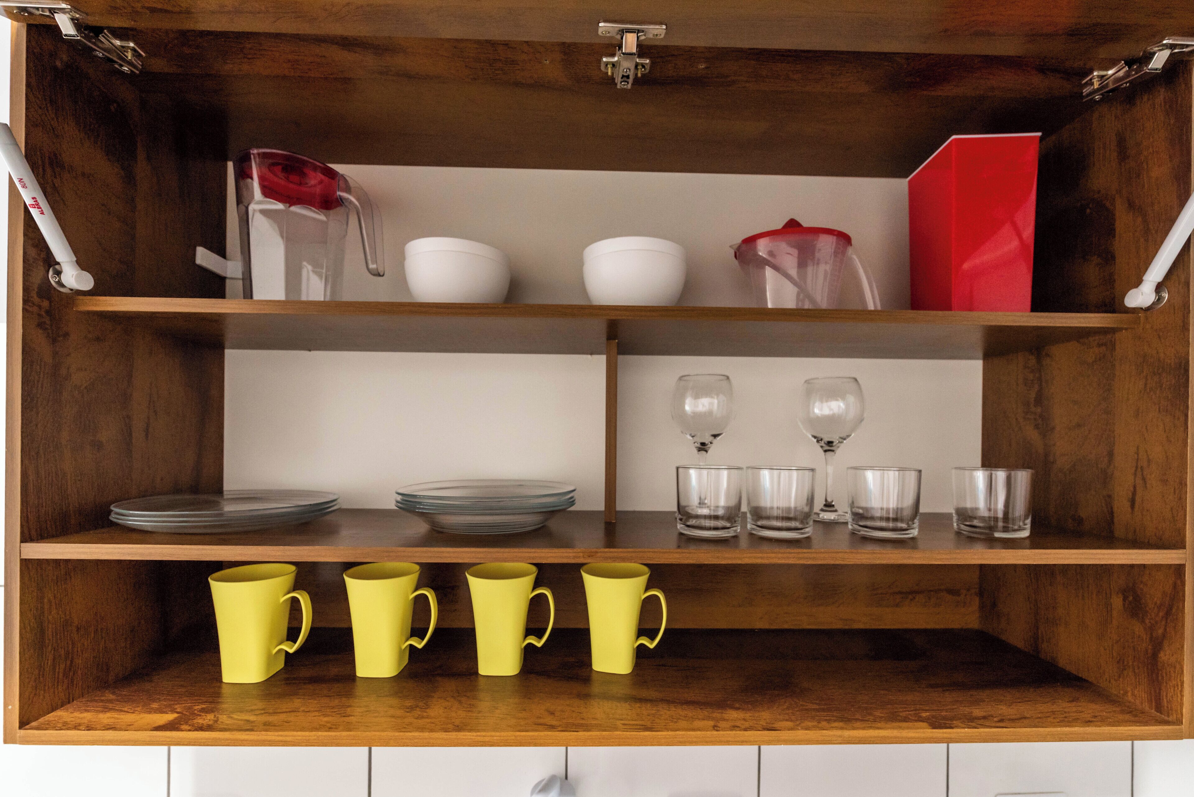 Una cocina con repisas de madera, tazas amarillas, tazones blancos y vasos transparentes.