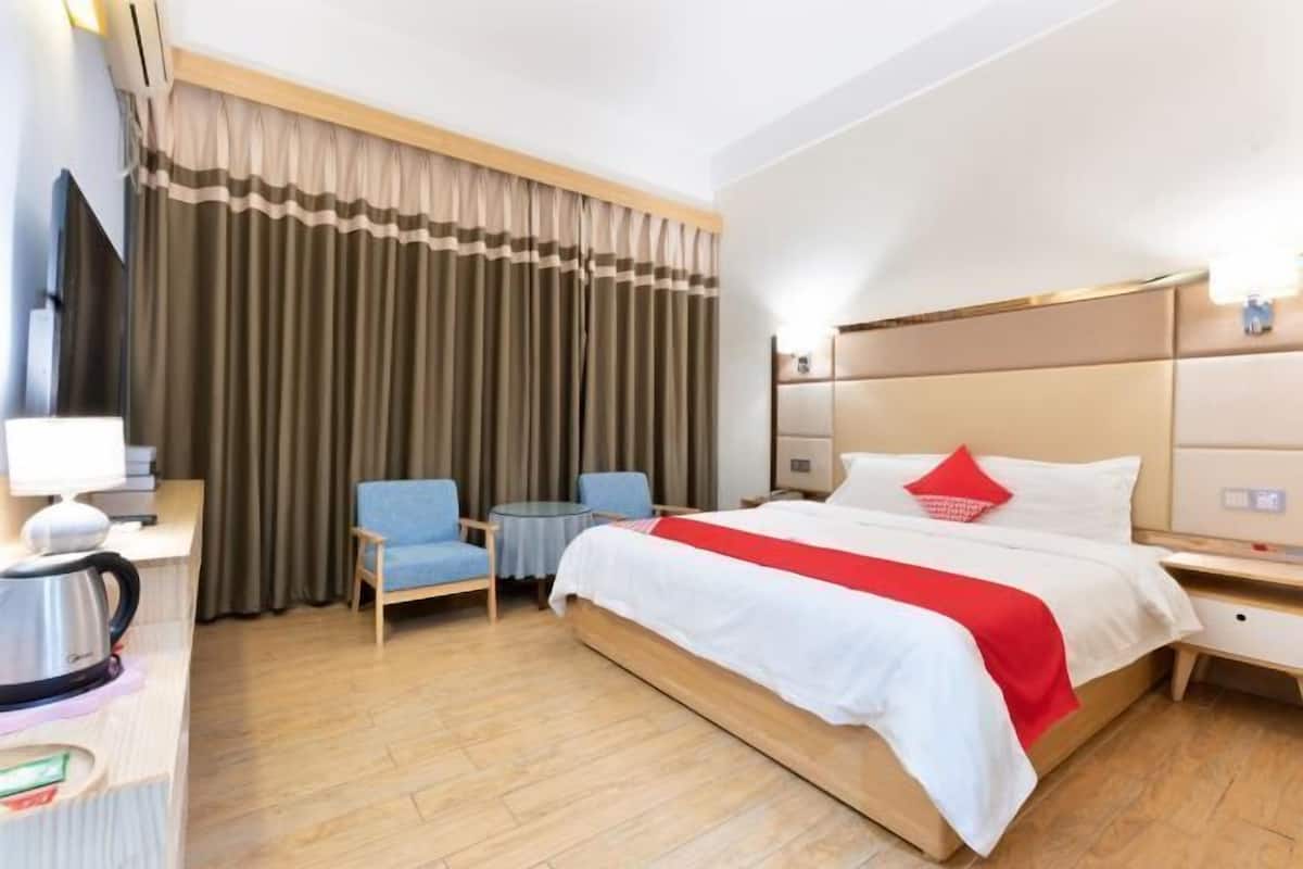 A hotel room with a bed, two chairs, a desk, a kettle, and a window with curtains.