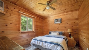 A wooden cabin bedroom with a bed, a nightstand, a lamp, a window, and a ceiling fan.