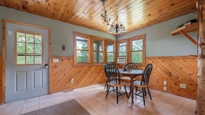 A dining area with a wooden table and chairs, a chandelier, and large windows with wooden frames.