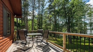 A wooden deck with a table and chairs, surrounded by a railing, overlooking a forested area and a body of water.