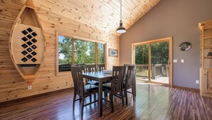 A wooden cabin interior with a dining table and chairs, large windows, and a wooden ceiling.