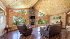 A spacious living area with wooden walls and a stone fireplace, large windows offering views of the outdoors.